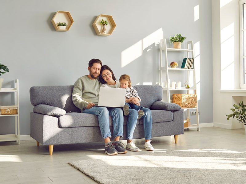 Full length photo of a happy young family with little child girl sitting on the sofa using modern laptop together. Parents resting on couch watching funny cartoon online or talking on video call.